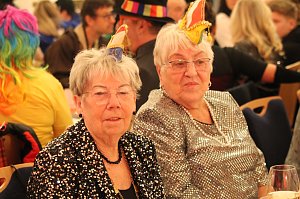 Karnevalsspektakel in einem gro&szlig;en Raum. Viele bunt gekleidete Menschen an Tischen und auf der B&uuml;hne