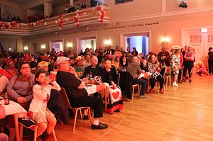 Karnevalsspektakel in einem gro&szlig;en Raum. Viele bunt gekleidete Menschen an Tischen und auf der B&uuml;hne